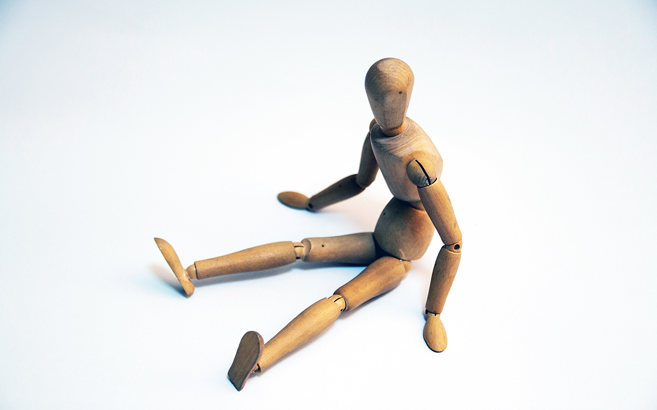 Wooden artist's mannequin seated with legs straight out, arms to the side, bracing, on a white surface