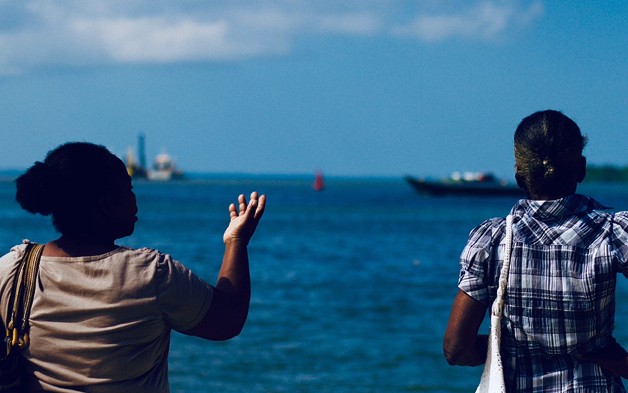 Two people seen from behind looking onto a waterway with a small boat in the distance.